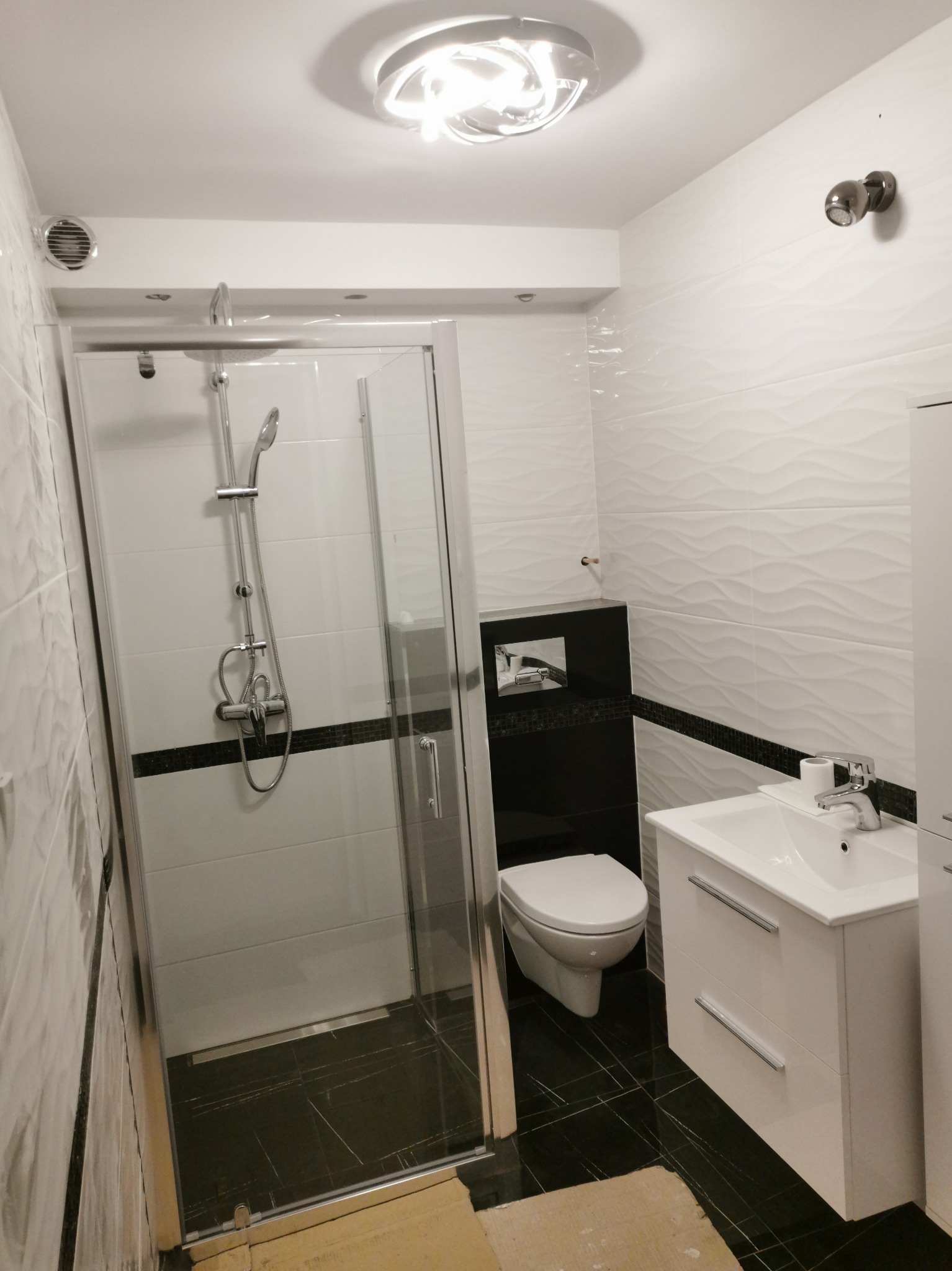 Nowoczesna łazienka z kabiną prysznicową, umywalką z szafką i podwieszaną toaletą, wykończona białymi płytkami o falistej strukturze i czarnymi akcentami.