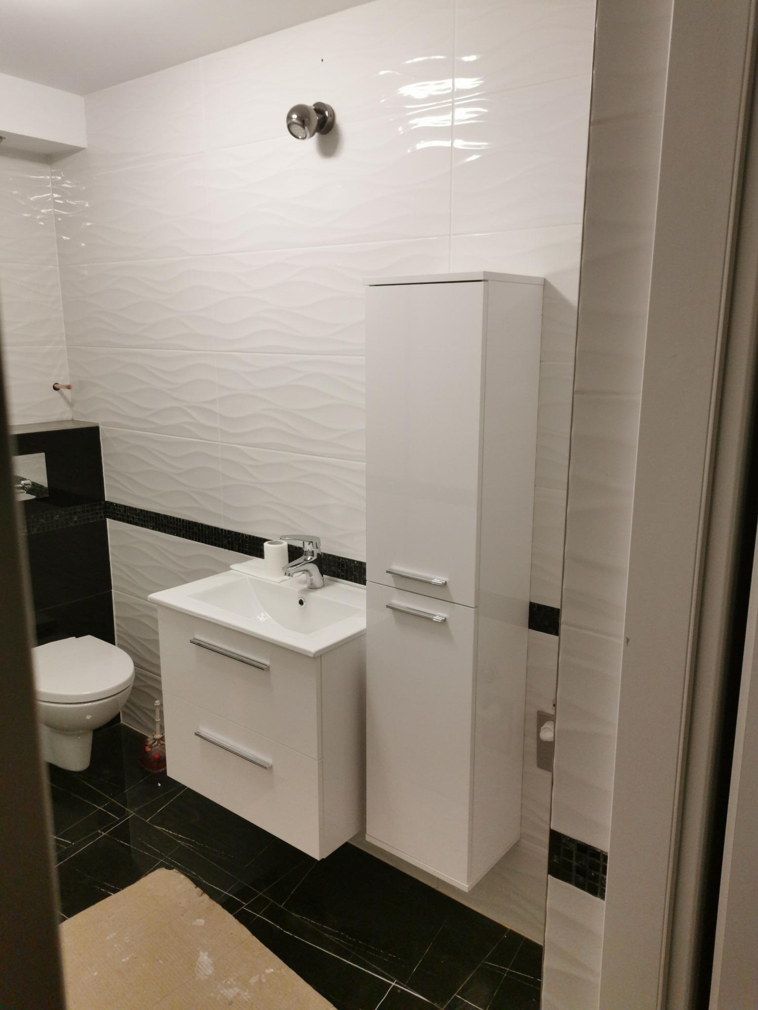 Nowoczesna łazienka z białymi, falistymi płytkami na ścianach, czarną listwą dekoracyjną, czarną podłogą z białymi żyłami, białą szafką pod umywalką i wysoką białą szafką łazienkową