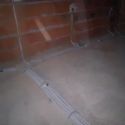 Surowe ściany z czerwonej cegły z widocznymi przewodami elektrycznymi ułożonymi na podłodze i wyprowadzonymi ze ściany, w trakcie prac instalacyjnych.