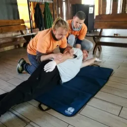 Instruktorzy w pomarańczowych koszulkach demonstrują na leżącym mężczyźnie techniki stabilizacji ramienia podczas szkolenia z pierwszej pomocy w sali z drewnianą podłogą i ławkami.