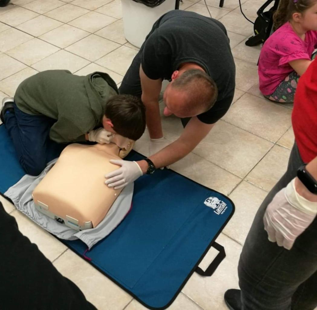 Uczeń wykonuje resuscytację usta-usta na manekinie pod okiem instruktora w białych rękawiczkach, leżącym na niebieskiej macie Laerdal.