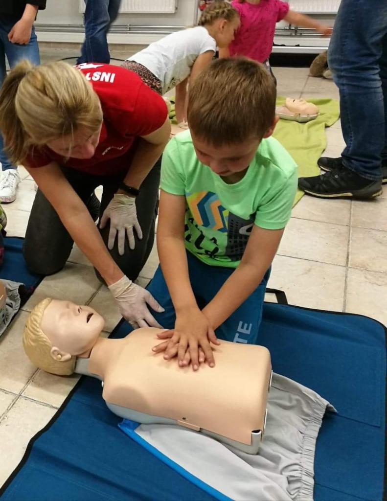 Instruktorka w rękawiczkach pokazuje chłopcu na manekinie, jak prawidłowo wykonywać resuscytację krążeniowo-oddechową podczas zajęć.