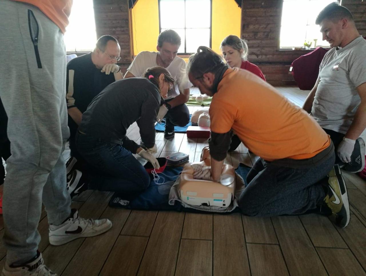 Grupa osób ćwiczy resuscytację krążeniowo-oddechową na manekinie, używając rękawic ochronnych, w pomieszczeniu z drewnianą podłogą i oknami w tle.