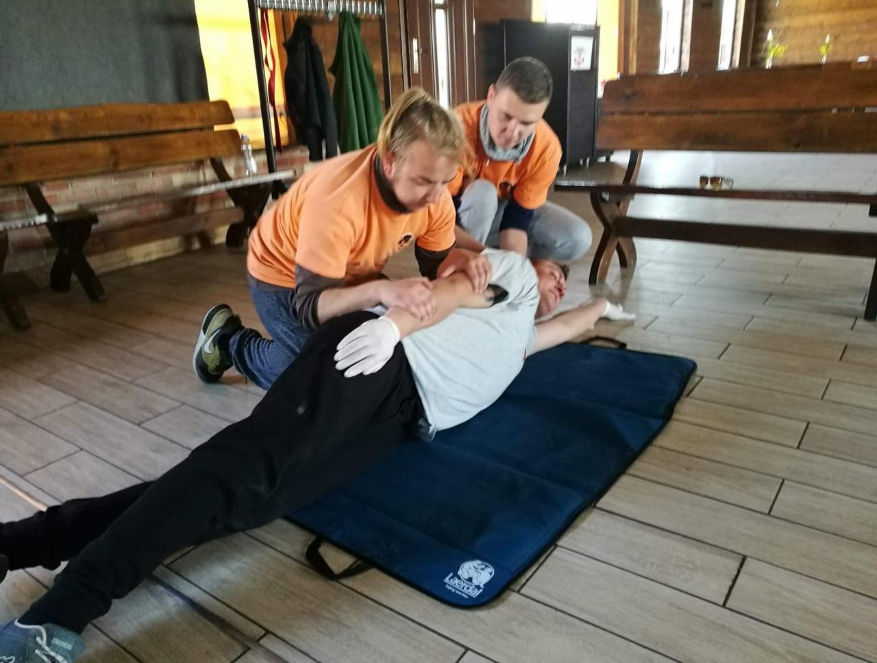 Instruktorzy w pomarańczowych koszulkach demonstrują na leżącym mężczyźnie techniki stabilizacji ramienia podczas szkolenia z pierwszej pomocy w sali z drewnianą podłogą i ławkami.