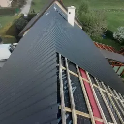 Montaż blachy trapezowej na dachu domu jednorodzinnego, widoczne łaty, kontrłaty oraz membrana dachowa, w tle zabudowa mieszkalna i zieleń.