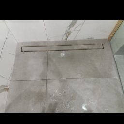 Express Team - Ukończony odpływ liniowy w kabinie prysznicowej, szare płytki podłogowe i marmurowe płytki ścienne, widoczne ślady wody po montażu.