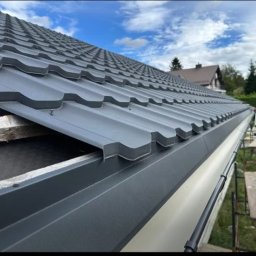Express Team - Szary metalowy dach z nowymi rynnami, widok z bliska na fragment dachu w trakcie montażu, z pracownikiem w tle i fragmentem budynku mieszkalnego w oddali.