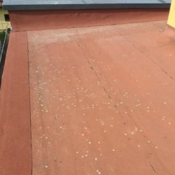 Płaski dach pokryty czerwoną papą z widocznymi śladami białych zabrudzeń i pęknięć, z fragmentem orynnowania i budynków w tle.