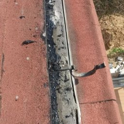 Express Team - Fragment dachu z czerwoną dachówką, widoczny brudny, uszczelniony na czarno element łączenia dachu z rynną, z metalowym uchwytem, w tle drzewa i gruz.