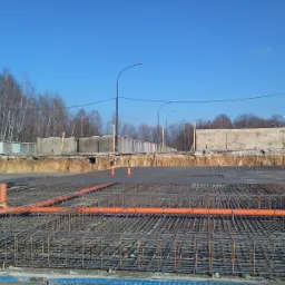 Zbrojenie fundamentów z rurami kanalizacyjnymi na tle wykopu i betonowych segmentów ogrodzenia pod błękitnym niebem.