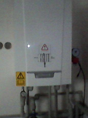 Biały piec grzewczy z widocznymi rurami, ostrzeżeniami i wskaźnikiem na ścianie.