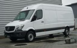 Biały samochód dostawczy Mercedes-Benz Sprinter zaparkowany na mokrym asfalcie przed szarą ścianą z blachy falistej. Widoczny lekki odblask na karoserii.