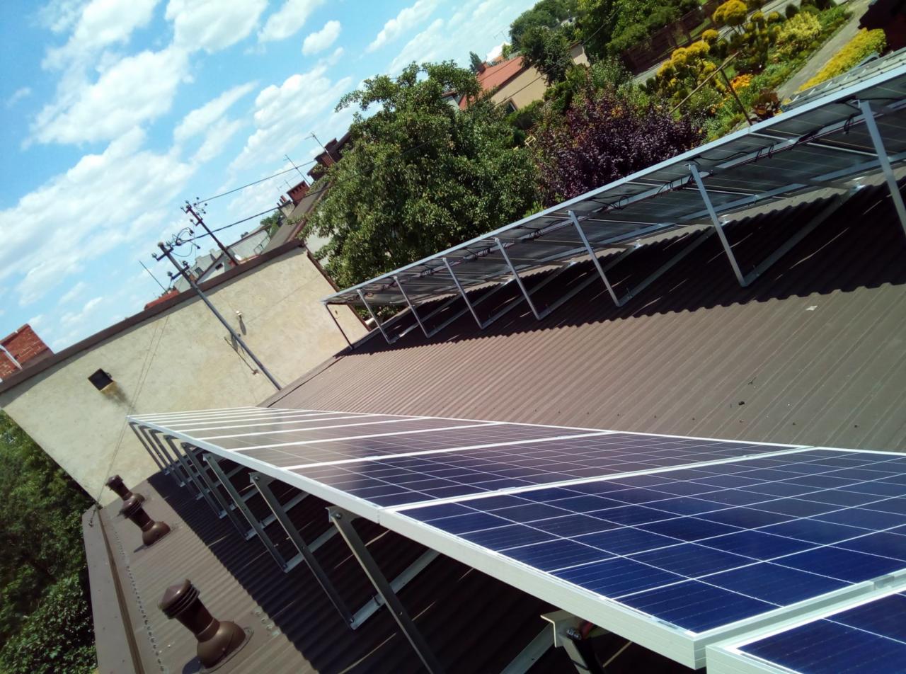 Panele słoneczne zamontowane na brązowym, falistym dachu budynku z widocznymi kominkami wentylacyjnymi, na tle błękitnego nieba z chmurami i zieleni drzew.