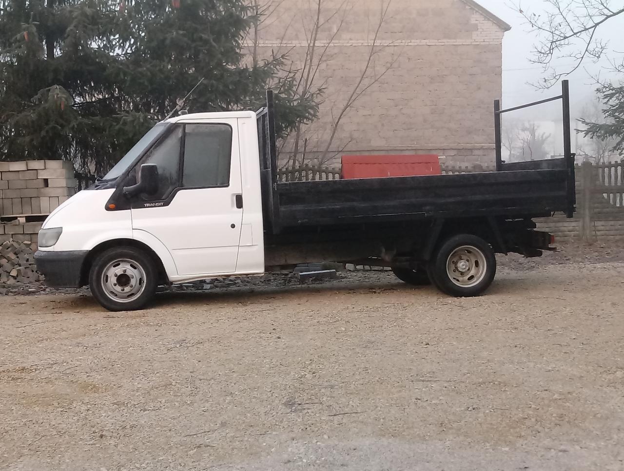 Biały Ford Transit z otwartą skrzynią ładunkową na żwirowym podłożu, z paletą kostki brukowej w tle, sfotografowany w pochmurny dzień.