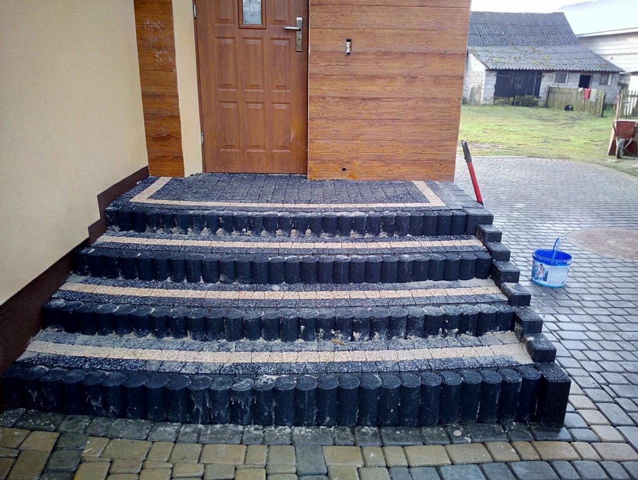 Czterostopniowe schody wejściowe wykończone czarną kostką imitującą drewno i jasnym żwirem, prowadzące do drzwi domu z drewnianą okładziną, z widocznym wiaderkiem i częścią posesji w tle.