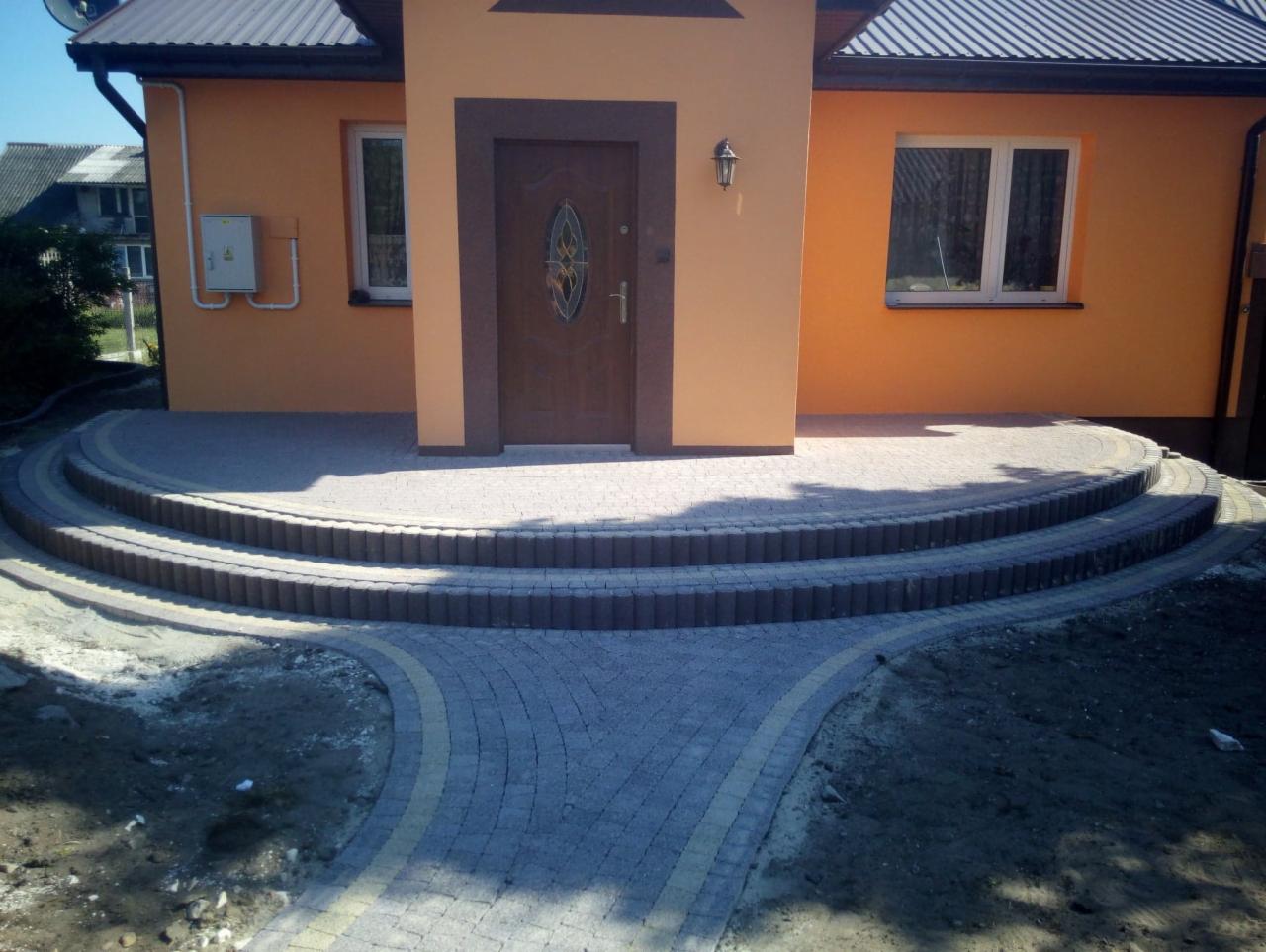 Ułożona kostka brukowa przed wejściem do domu z trzema półokrągłymi stopniami, widoczne świeże ślady po pracach ziemnych.