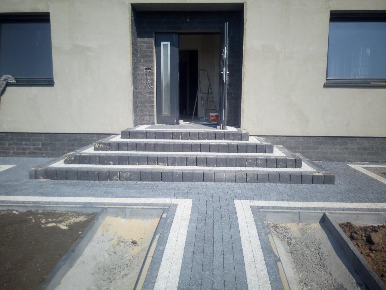 Schody wejściowe i chodnik z kostki brukowej w odcieniach szarości i bieli, z widocznymi fragmentami niedokończonych rabat po bokach, na tle surowej elewacji budynku z otwartymi drzwiami.