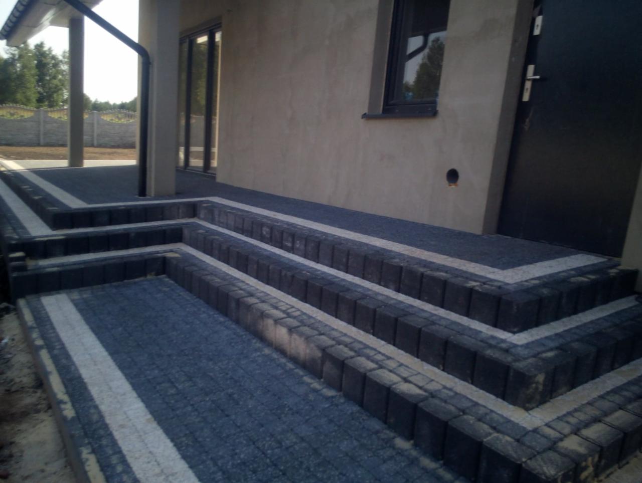 Schody wejściowe do domu wyłożone ciemną kostką brukową z jasnymi pasami dekoracyjnymi, prowadzące do drzwi i okna.