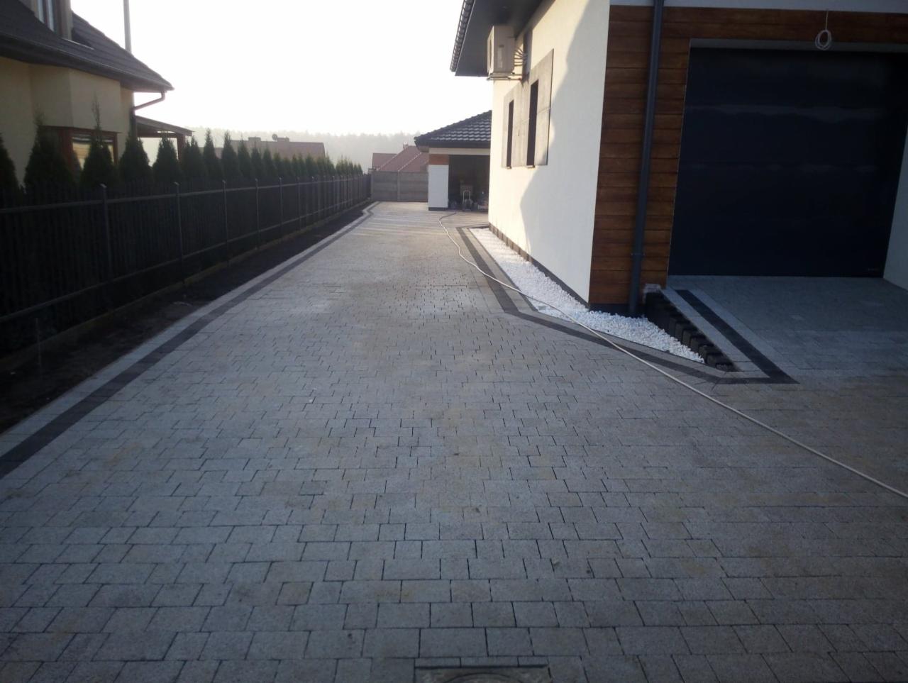 Ułożona szara kostka brukowa na podjeździe do domu jednorodzinnego z czarną bramą garażową i białym kamieniem ozdobnym wzdłuż ściany.