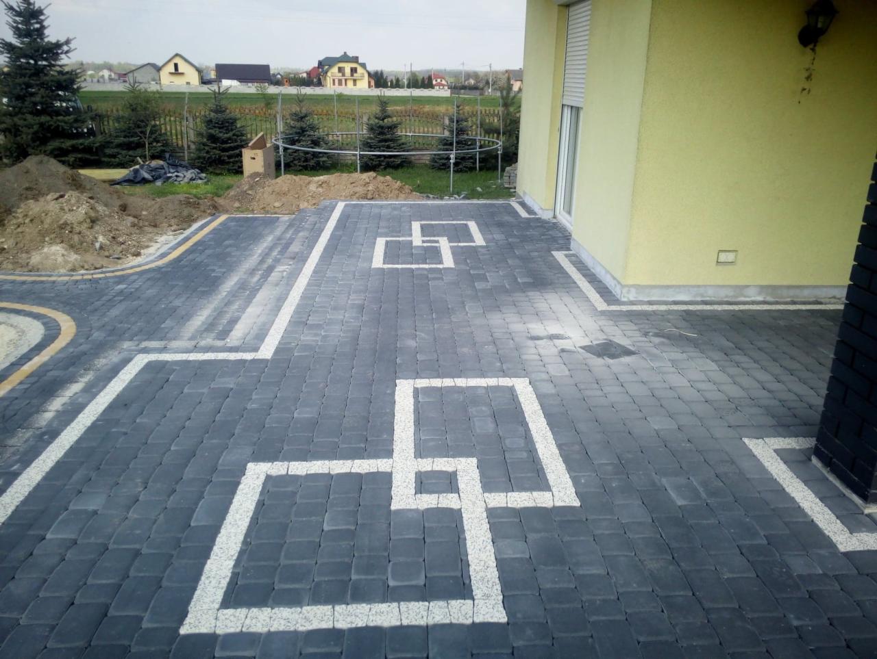 Ułożona szara kostka brukowa z dekoracyjnymi białymi wstawkami tworzącymi geometryczne wzory, widoczny fragment elewacji budynku i przygotowany teren pod nasadzenia.