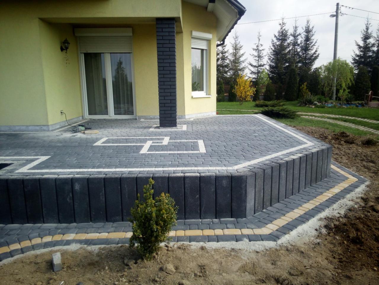 Wykończony taras z kostki brukowej przed domem o żółtej elewacji, z obrzeżem z ciemnych palisad i jasną fugą, wzór z białych kostek na powierzchni tarasu, widoczne elementy ogrodu w tle.