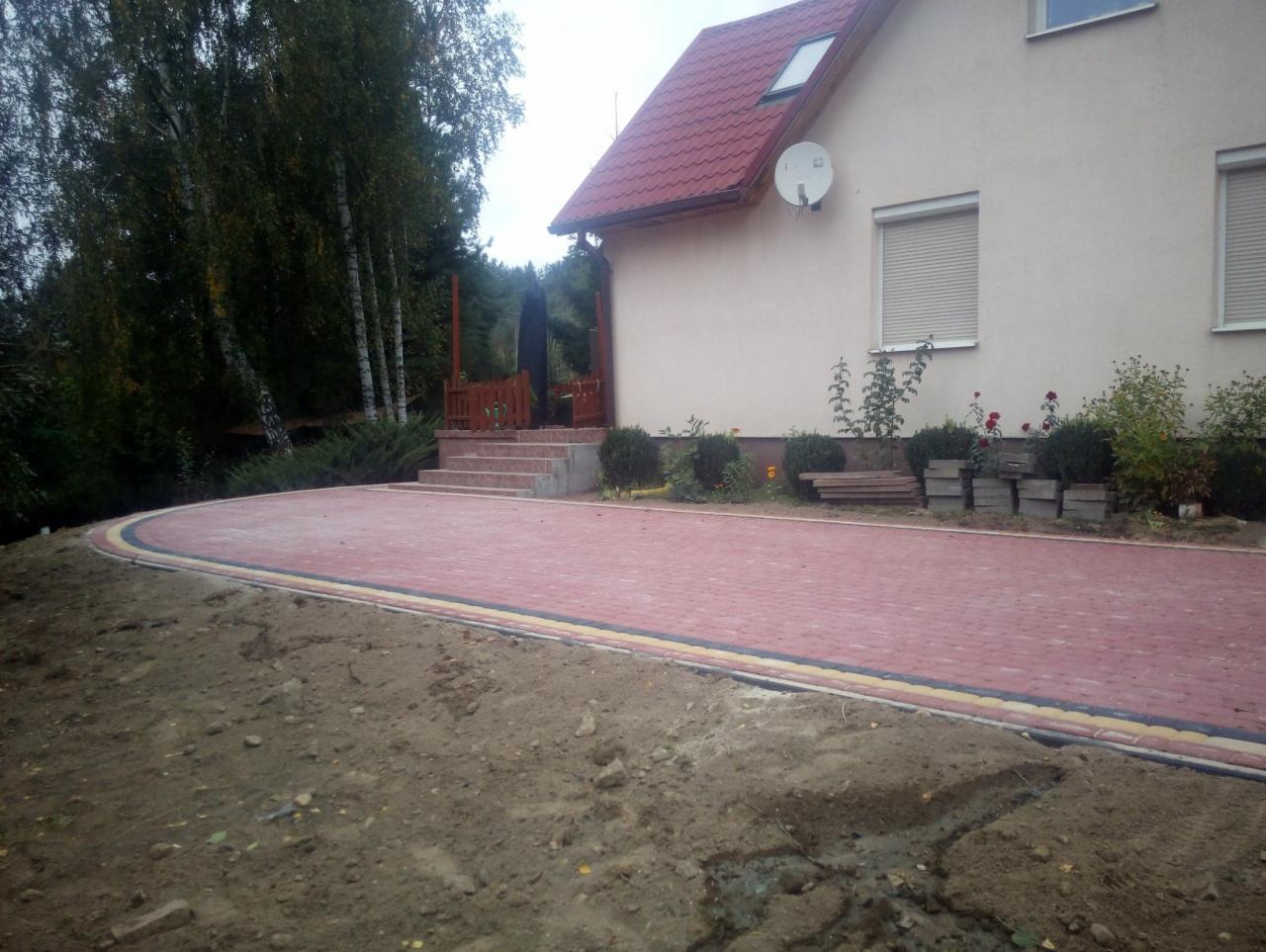 Czerwona kostka brukowa ułożona na posesji przed domem jednorodzinnym z widocznymi elementami krajobrazu i schodami wejściowymi.