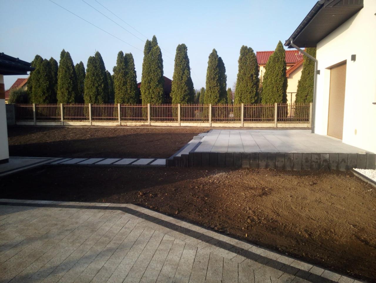 Nowoczesny taras z szarej kostki brukowej, z obrzeżem z ciemnych bloczków, prowadzący do domu, z widocznym fragmentem ścieżki z płyt betonowych w ogrodzie, w tle rząd tuj i ogrodzenie.
