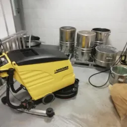 Żółta maszyna czyszcząca Karcher na szarym, lekko wilgotnym podłożu, w tle białe kafelki i metalowe pojemniki.