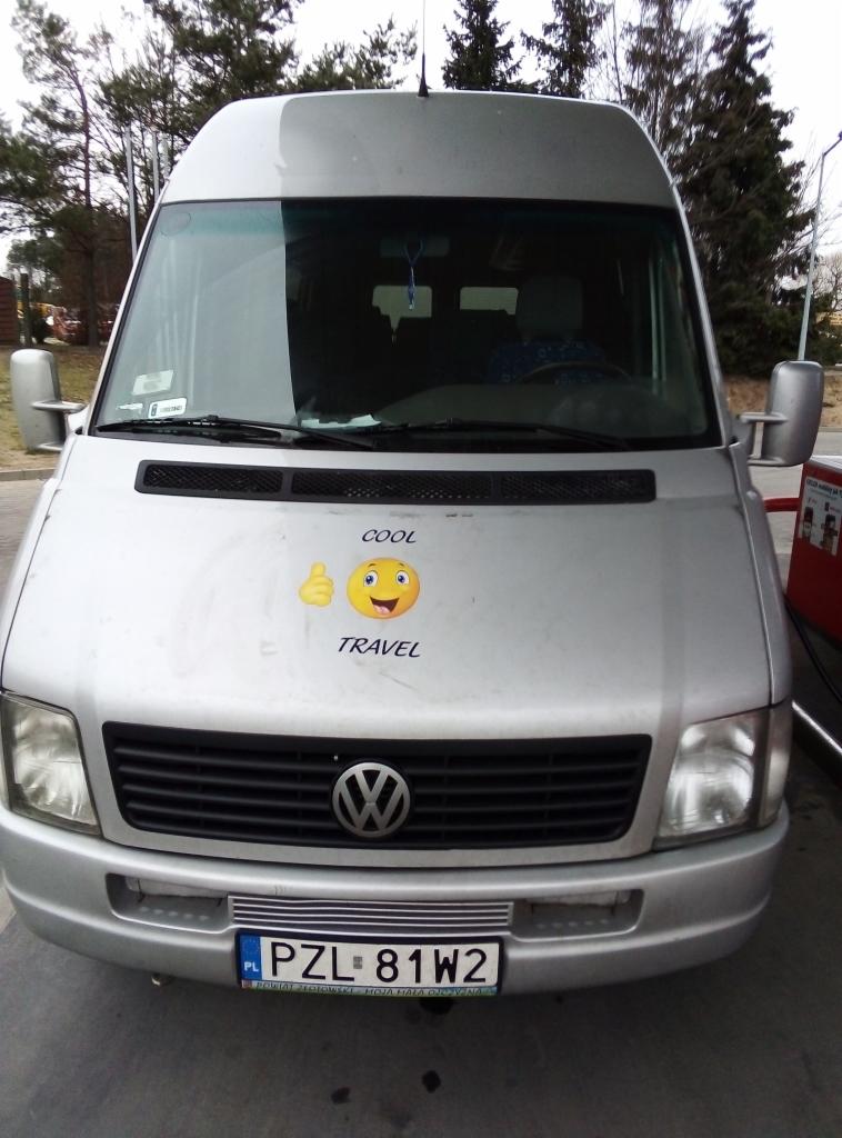 Srebrny bus Volkswagen z naklejką 'COOL TRAVEL' na masce, zaparkowany na zewnątrz, widok z przodu.