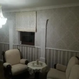 Elegancki pokój po remoncie: tapeta w ornamenty, kolumny dekoracyjne, kremowe fotele i kryształowy żyrandol.