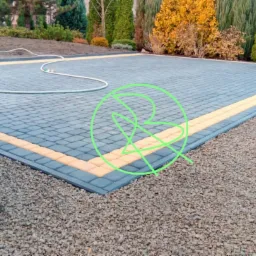 Nowo ułożony podjazd z szarej kostki brukowej z żółtym obramowaniem, otoczony kamiennym grysem i zielenią ogrodu, widoczny wąż ogrodowy.