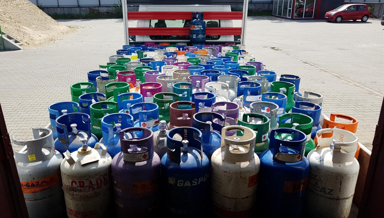 Wnętrze samochodu dostawczego wypełnione butlami gazowymi w różnych kolorach, ułożonymi rzędami, z pojedynczą butlą BUTAN widoczną w tle, na zewnątrz widoczny plac z kostki brukowej i budynek.