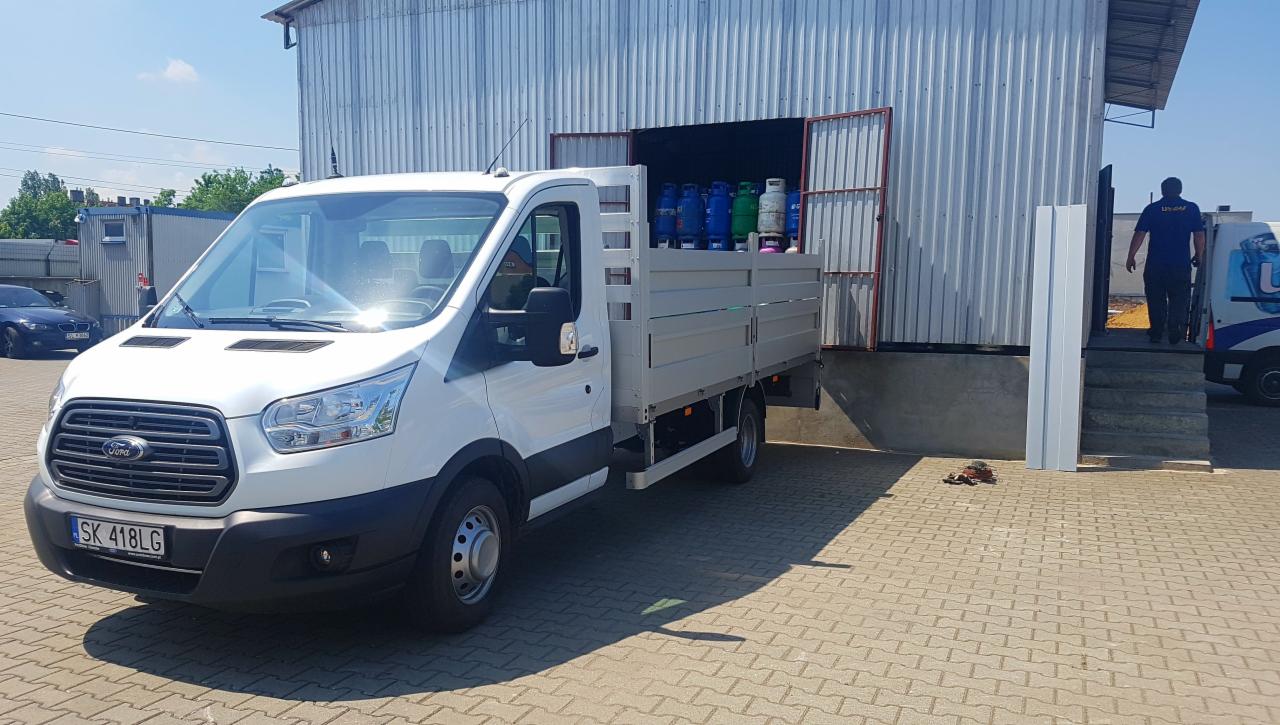 Biały samochód dostawczy Ford z otwartą skrzynią ładunkową, wypełnioną butlami gazowymi w różnych kolorach, zaparkowany przed magazynem z otwartymi drzwiami, z widocznym pracownikiem wchodzącym...