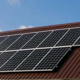 Panele słoneczne zamontowane na brązowym dachu z blachy trapezowej pod błękitnym niebem z lekkimi chmurami.