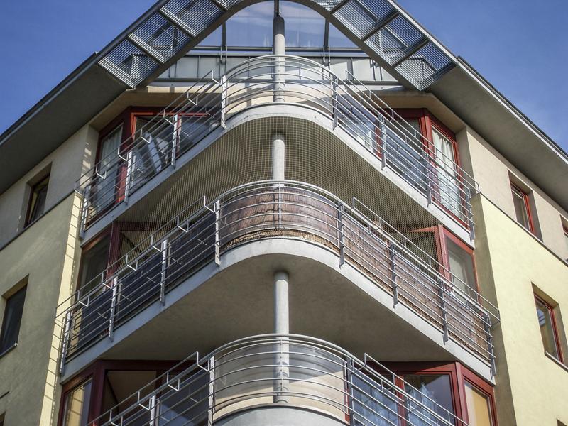 Fasada budynku z narożnymi balkonami zabezpieczonymi siatką ochronną przed ptakami, widok od dołu na trzy kondygnacje.