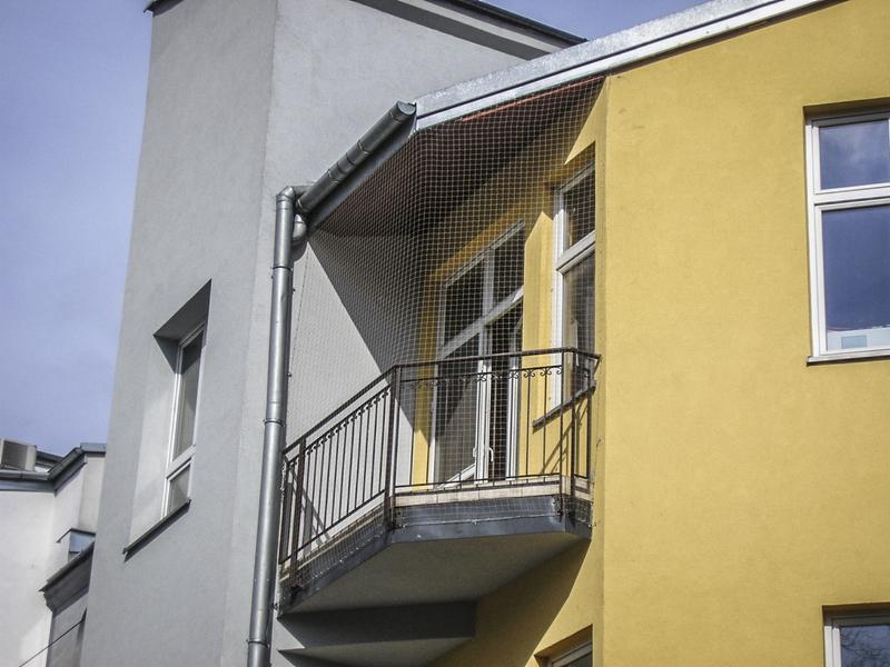 Zabezpieczony balkon siatką ochronną przed ptakami, zamontowaną na metalowej konstrukcji, widok z dołu na żółto-szary budynek.