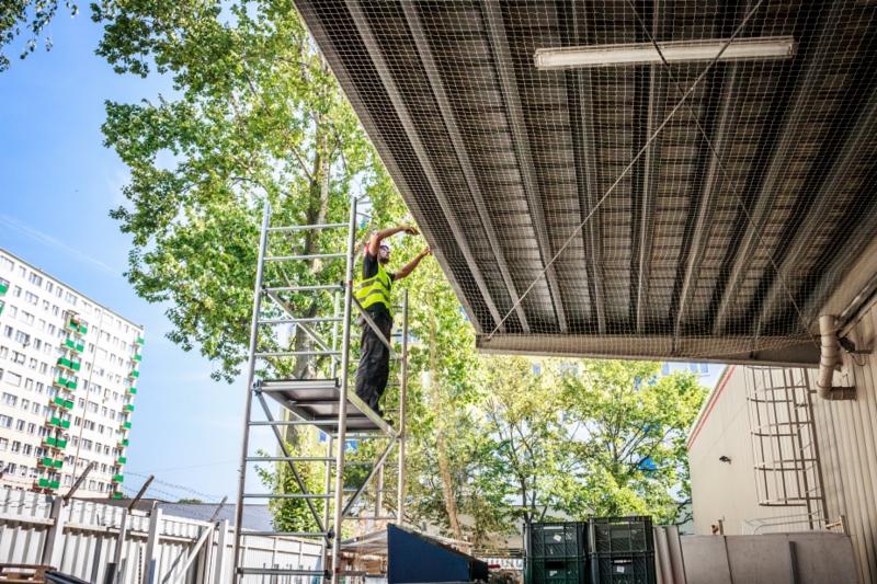 Pracownik w kamizelce odblaskowej montuje siatkę zabezpieczającą pod zadaszeniem, stojąc na aluminiowym rusztowaniu na tle drzew i budynku mieszkalnego.