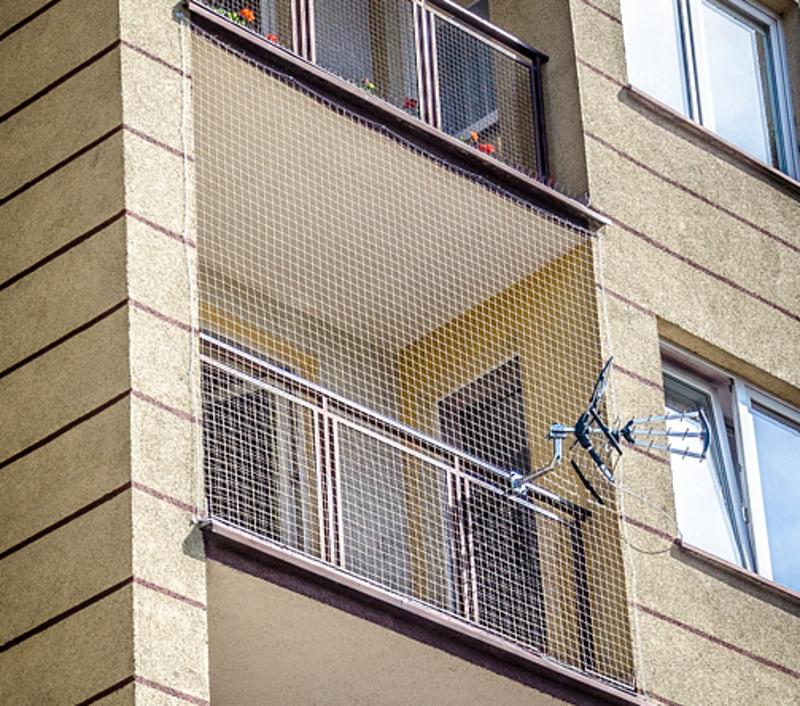 Zabezpieczony siatką balkon w bloku z widoczną anteną satelitarną, kwiaty w doniczkach na górnej krawędzi balkonu.