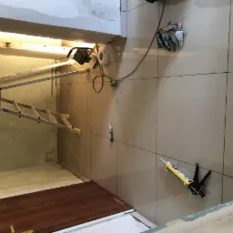 Wnętrze w trakcie remontu: drabina przystawiona do ściany z widocznymi śladami po tynkowaniu, podłoga z płytek ceramicznych, narzędzia i akcesoria budowlane w tle, lampa robocza na suficie, ściana...