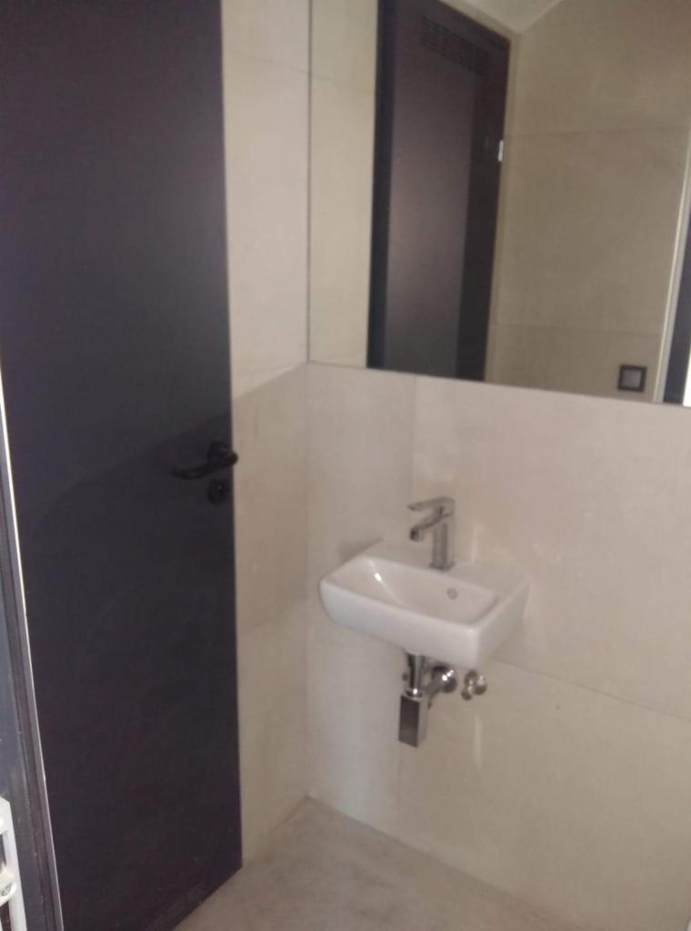 Mała, biała umywalka zamontowana w rogu łazienki w Warszawie, z widocznym lustrem i ciemnymi drzwiami w tle.