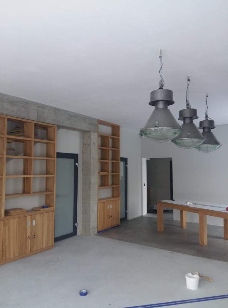 Wnętrze w trakcie wykańczania: surowy betonowy filar, drewniane regały z szafkami bez frontów, trzy industrialne lampy wiszące, zabezpieczony stół, podłoga wylewka, przygotowane do malowania ściany.