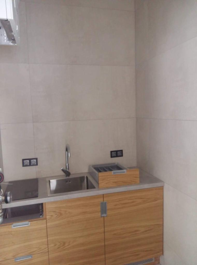 Wykończony fragment kuchni z drewnianymi szafkami, stalowym zlewem i baterią, oraz beżowymi płytkami na ścianie. Nad blatem widoczne gniazdka elektryczne.