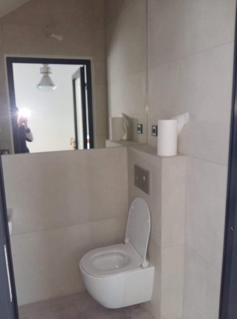 Nowoczesna, podwieszana toaleta z otwartą klapą w wykończonej łazience, widoczne płytki w odcieniach beżu i szarości, odbicie fotografa w lustrze.