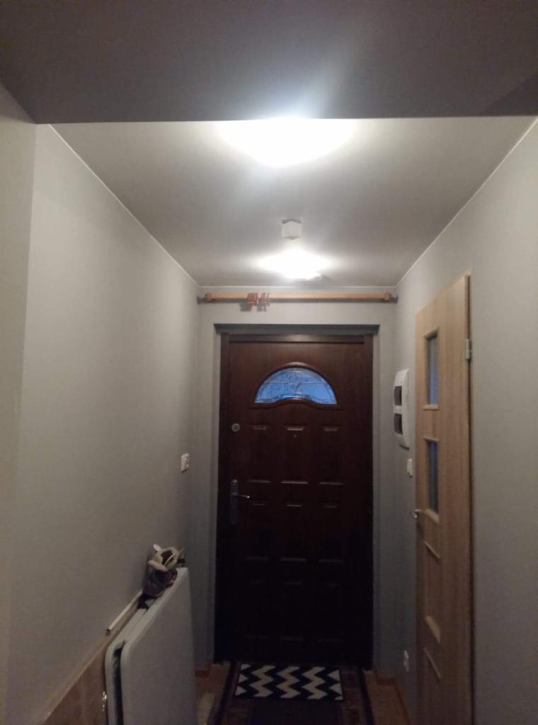 Przedpokój po remoncie: szare ściany, biały sufit z lampami, ciemne drzwi wejściowe z półokrągłym okienkiem, jasne drzwi wewnętrzne, rura pod sufitem, składany stół przy ścianie.