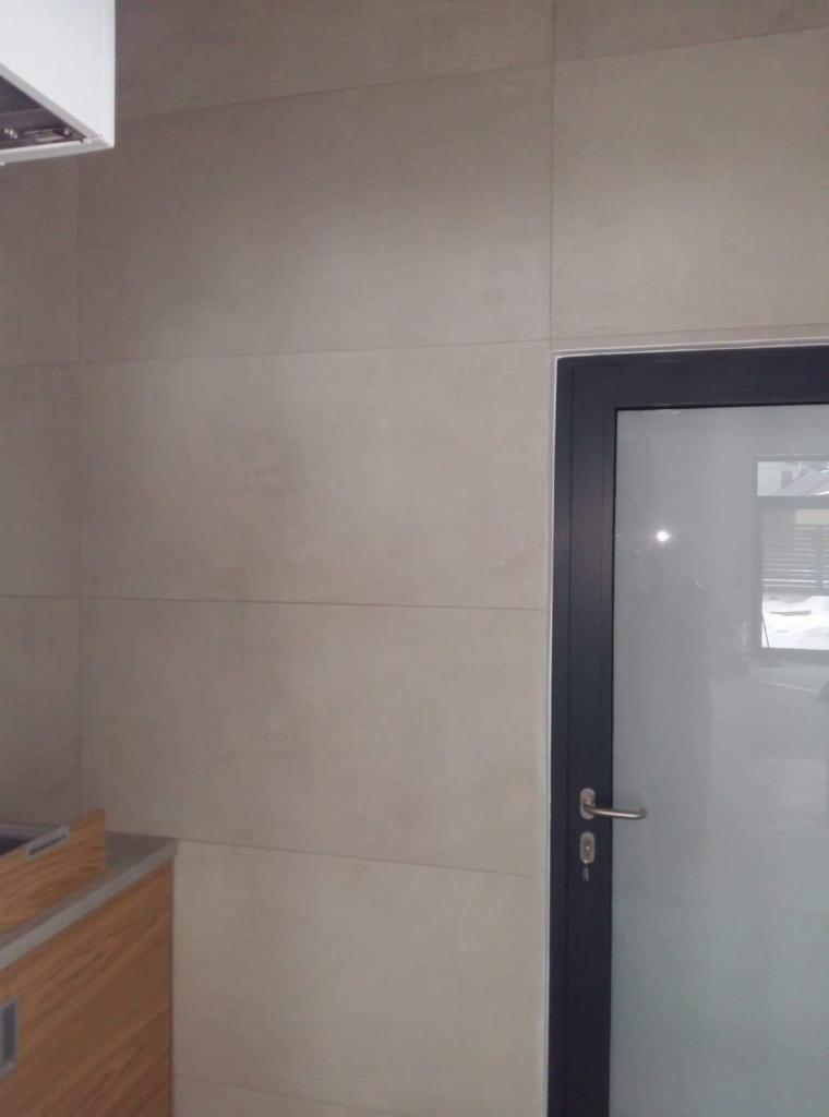 Beżowe, matowe płytki wielkoformatowe na ścianie w łazience, widoczna szara rama szklanych drzwi oraz fragment szafki pod umywalkę.