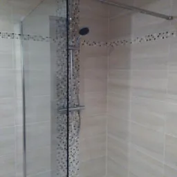 Kabina prysznicowa z deszczownicą i mozaiką z drobnych płytek w beżowych odcieniach, zamontowana na jasnych, podłużnych płytkach ściennych.