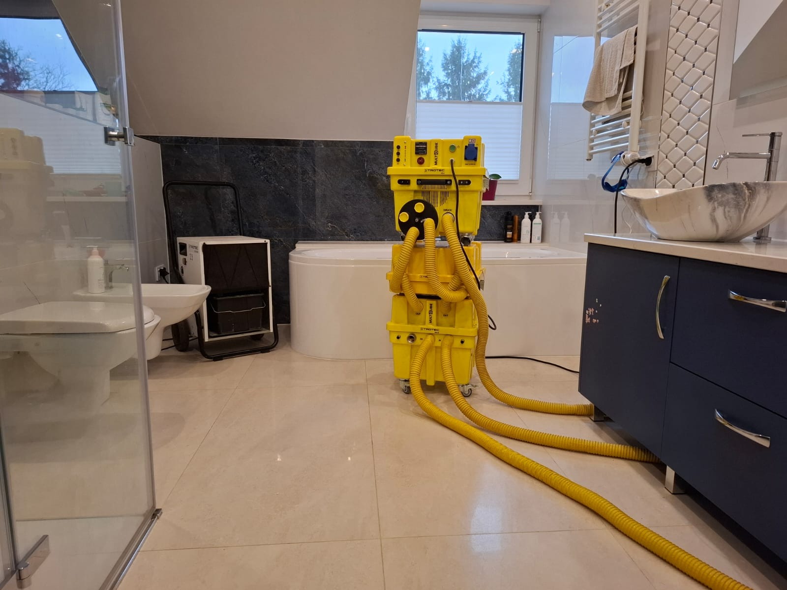 Żółty, modułowy osuszacz podposadzkowy Trotec MultiQube w łazience z beżowymi płytkami, widoczne węże i elementy sterujące, w tle wanna i umywalka.