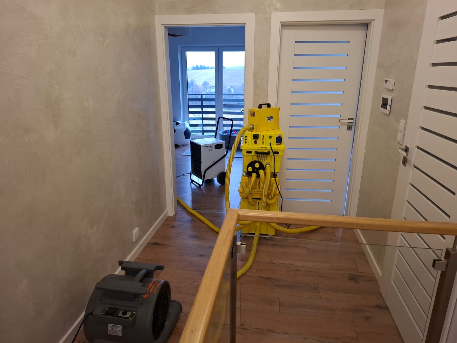 Profesjonalny sprzęt do osuszania w trakcie pracy w korytarzu z drewnianą podłogą, widoczny osuszacz kondensacyjny i wentylator osuszający.