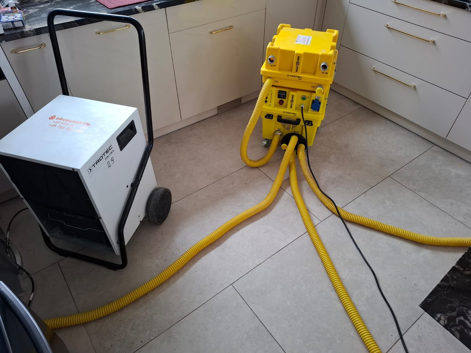 Dwa urządzenia do osuszania powietrza marki Trotec, jeden biały model TTK 170 S z kółkami i uchwytem, drugi żółty Multicube, połączone żółtymi wężami, ustawione na jasnych płytkach podłogowych...
