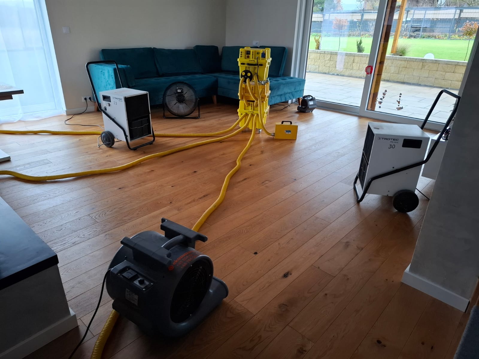 Zestaw urządzeń do usuwania wilgoci w pomieszczeniu z parkietem, widoczny narożnik turkusowej sofy i przesuwne drzwi tarasowe